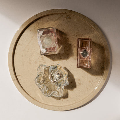 Kunstvolle Ambient-Aufnahme des runden Travertin-Tabletts von Marmonix, edel inszeniert mit exklusiven Parfümflakons und eleganten Licht- und Schattenspielen auf einem modernen Frisiertisch.