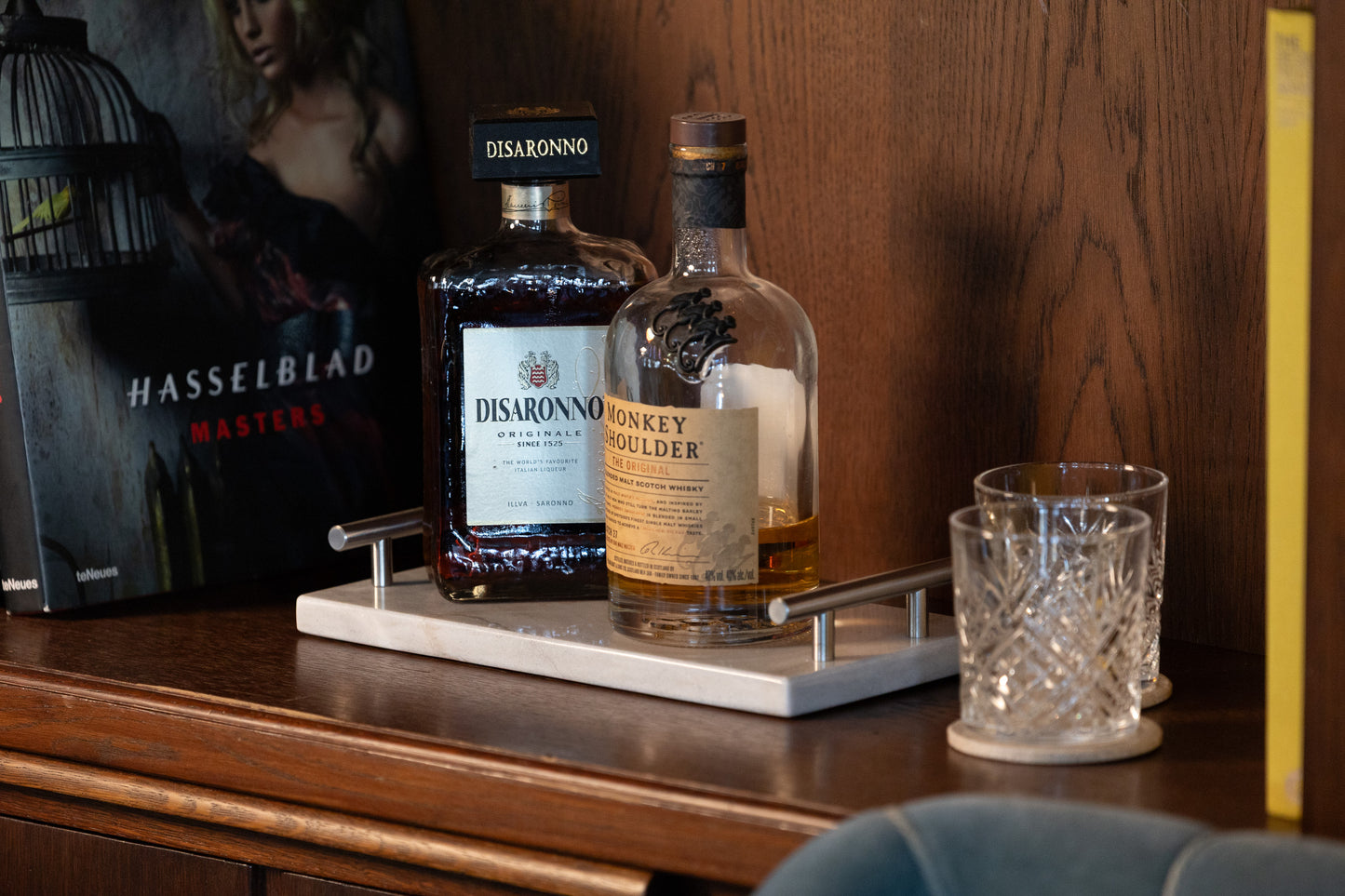 Stimmungsvolle Ambient-Aufnahme des weißen Marmortabletts mit Griffen von Marmonix, exklusiv platziert in einem Bücherregal mit zwei Gläsern und edlem Whiskey als stilvolle Heimbar-Inszenierung.